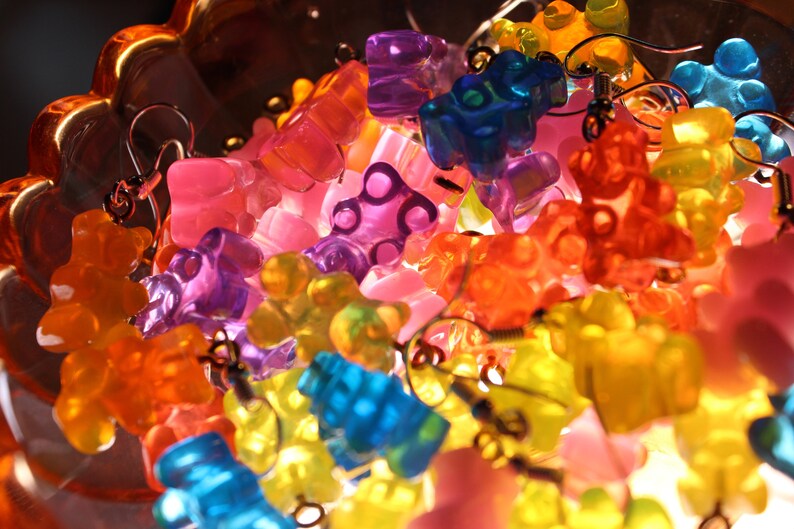 Pu&ograve; includere: Orecchini a forma di orsetti gommosi colorati in varie tonalit&agrave; di arancione, giallo, rosa, viola e blu. Gli orecchini sono in plastica traslucida e hanno ganci color argento. Gli orecchini sono esposti in una ciotola di vetro decorativa.
