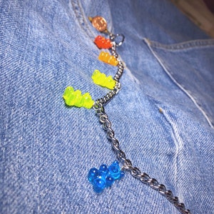 Pu&ograve; includere: Una catena d'argento con ciondoli a forma di orsetti gommosi colorati, tra cui rosso, arancione, giallo e blu. La catena &egrave; attaccata a una tasca di jeans, con una piccola moneta visibile in alto.