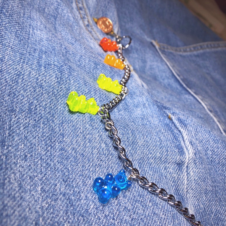Pu&ograve; includere: Una catena d'argento con ciondoli a forma di orsetti gommosi colorati, tra cui rosso, arancione, giallo e blu. La catena &egrave; attaccata a una tasca di jeans, con una piccola moneta visibile in alto.