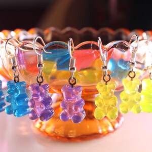 Pu&ograve; includere: Orecchini a forma di orsetti gommosi colorati in giallo, blu, viola e verde, appesi a ganci d'argento. Gli orecchini sono esposti in una ciotola di vetro decorativa con una tonalit&agrave; arancione e arcobaleno.