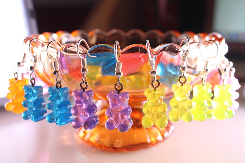 Pu&ograve; includere: Orecchini a forma di orsetti gommosi colorati in giallo, blu, viola e verde, appesi a ganci d'argento. Gli orecchini sono esposti in una ciotola di vetro decorativa con una tonalit&agrave; arancione e arcobaleno.