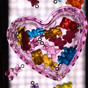 Pu&ograve; includere: Un piatto di vetro rosa a forma di cuore pieno di ciondoli a forma di orsetti gommosi colorati. I ciondoli sono in vari colori, tra cui rosso, giallo, blu, arancione e viola. Alcuni ciondoli sono fuori dal piatto.