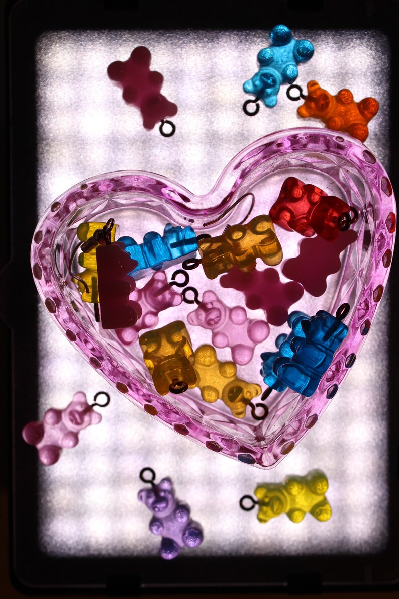 Pu&ograve; includere: Un piatto di vetro rosa a forma di cuore pieno di ciondoli a forma di orsetti gommosi colorati. I ciondoli sono in vari colori, tra cui rosso, giallo, blu, arancione e viola. Alcuni ciondoli sono fuori dal piatto.