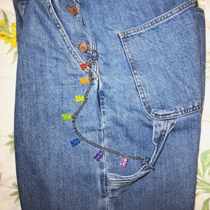Pu&ograve; includere: Primo piano di jeans blu con una catena decorativa. La catena &egrave; adornata da ciondoli a forma di orsetti gommosi colorati nelle tonalit&agrave; del rosso, arancione, giallo, verde, blu e viola. I jeans hanno un classico design con bottoni e tasche.