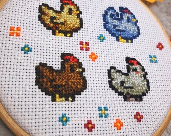 以下が含まれることがあります： 木製のフレームに収められたクロスステッチ刺繍。茶色、青、グレーの色合いのピクセル化された鶏が4羽おり、それぞれ赤い鶏冠が付いています。デザインには、小さくカラフルな花のアクセントが含まれています。