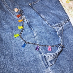 Pu&ograve; includere: Primo piano di jeans blu con una catena d'argento e ciondoli a forma di orsetti gommosi colorati. La catena &egrave; attaccata al bottone e alla tasca dei jeans. Gli orsetti gommosi sono rossi, arancioni, gialli, verdi, blu, viola e rosa.