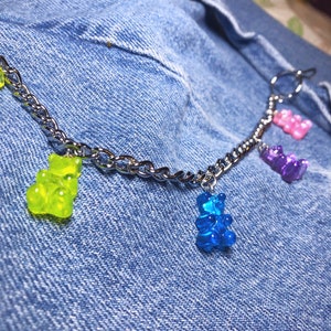 Pu&ograve; includere: Una collana a catena argentata con ciondoli a forma di orsetti gommosi verdi, blu, viola e rosa. La collana &egrave; presentata su uno sfondo in denim, che mette in risalto i colori vivaci dei ciondoli.