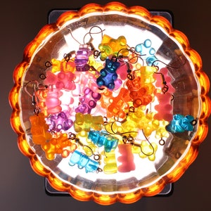 Pu&ograve; includere: Una collezione di orecchini a forma di orsetti gommosi colorati in varie tonalit&agrave; di rosa, arancione, giallo, blu e viola. Gli orecchini sono esposti in un piatto trasparente con bordo smerlato e una luce arancione.