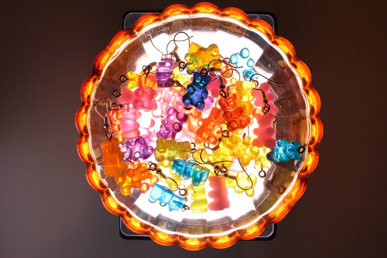 Pu&ograve; includere: Una collezione di orecchini a forma di orsetti gommosi colorati in varie tonalit&agrave; di rosa, arancione, giallo, blu e viola. Gli orecchini sono esposti in un piatto trasparente con bordo smerlato e una luce arancione.