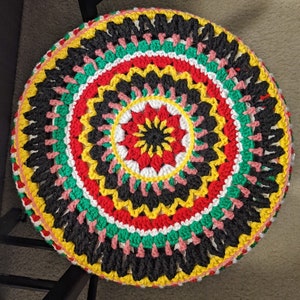 May include: A round, crocheted cushion with a vibrant, concentric design. The cushion features rings of black, red, yellow, green, and white, with a central red and yellow floral pattern. The edges are finished with pink and yellow yarn.