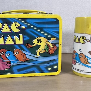 May include: A vintage yellow Pac-Man lunchbox and matching thermos. The lunchbox features the Pac-Man logo and characters on a blue background. The thermos has a yellow lid and a white body with Pac-Man graphics.
