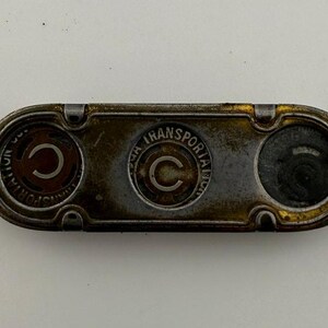 May include: Vintage metal token with the text "C" and "TRANSPORTATION CO." in a circular design. The token has a rectangular shape with rounded ends and a tarnished gold and brown color scheme. The token has three circular openings.