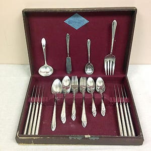 May include: A silver-toned flatware set arranged in a dark brown storage case with a deep red velvet interior. The set includes various spoons, forks, and knives, featuring a decorative pattern on the handles and a blue diamond-shaped label.
