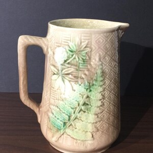 May include: Tan ceramic pitcher with a handle and spout. The pitcher features a raised diamond pattern and embossed green and white botanical designs, including ferns and other foliage. The pitcher is a decorative item.