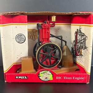 May include: A red and black IHC Titan Engine die-cast metal model inside a red and white box. The model features a large black flywheel, a gold-colored cylinder, and a brown base. The box has the text "Made in America" and "IHC Titan Engine".