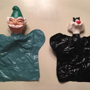 May include: Two vintage hand puppets. One puppet is a smiling gnome with a green hat and teal body. The other is a black cat puppet with a red nose and black and white face. Both puppets have a paper-like texture.