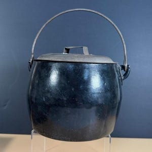 May include: A black, antique cast iron pot with a lid and a metal handle. The pot has a rounded shape and a slightly glossy finish. The handle arches over the top, and the lid has a small handle. The pot is likely used for cooking.