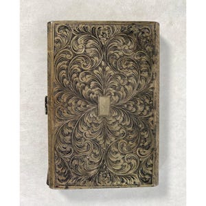 May include: A rectangular, antique-style box with an ornate, engraved floral pattern. The box is a muted gold color with dark accents in the engraved details. A small rectangular plate is centered on the front. The box has a clasp on the side.