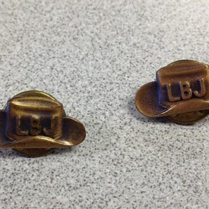 May include: Two bronze-colored cowboy hat pins with the letters "LBJ" on the front. The pins are detailed with a hat brim and crown, and are set against a speckled gray background.