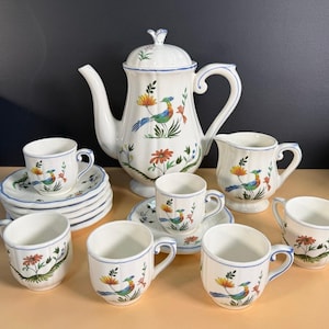 May include: A white ceramic tea set with a floral and bird design. The set includes a teapot, creamer, sugar bowl, saucers, and cups. The rims are trimmed with blue. The teapot has a lid with a decorative finial.
