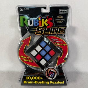 May include: Rubik's Slide electronic puzzle game in its original packaging. The game features a black and white grid with red and blue squares. The packaging includes the text "Ages 8 to 108" and "10,000+ Brain-Busting Puzzles!"