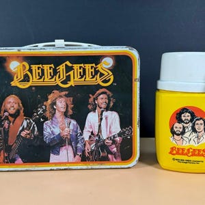 May include: A rectangular metal lunchbox featuring a graphic of the Bee Gees performing with guitars and the text Bee Gees, paired with a matching yellow plastic thermos featuring a circular portrait of the band and the text Bee Gees.