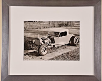 May include: A framed sepia-toned photograph of a custom hot rod car with an exposed engine, large rear tires, and a soft top. The image is encased in a wide, brushed metallic gray frame with a thick white mat border surrounding the print.