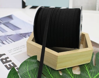 May include: A spool of black suede cord, wound around a black plastic bobbin, sitting on a wooden tray. The cord is lying on a green leaf.