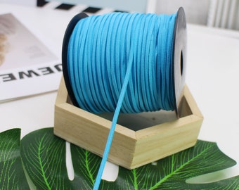 May include: A spool of bright turquoise suede cord sits in a small wooden box. The cord is tightly wound on a black spool. A single strand of the cord extends from the spool. The background includes a green leaf and a white surface.