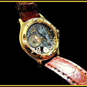 May include: A gold-tone wristwatch with a brown leather band. The watch face is clear and shows the inner workings of the watch.