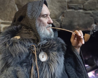 May include: A man wearing a gray hooded cloak with a fur collar and a leather strap. He is holding a wooden pipe in his mouth and looking to the right.