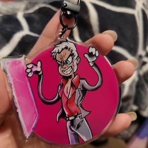 May include: A round pink and white acrylic keychain featuring a cartoon character with white hair, a red shirt, and a gray jacket. The character is making a funny face and has a pink background.