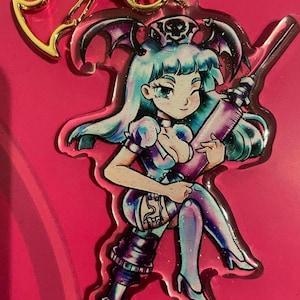 May include: A clear acrylic keychain featuring a cartoon nurse with blue hair, wearing a white dress and holding a large syringe. The nurse has bat wings and a skull on her hat. The keychain has a gold metal chain.