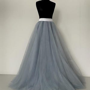 May include: A full-length, gray tulle skirt with a white waistband is displayed on a black mannequin. The skirt is voluminous and flowing, creating a soft, ethereal look. The fabric appears lightweight and semi-sheer.