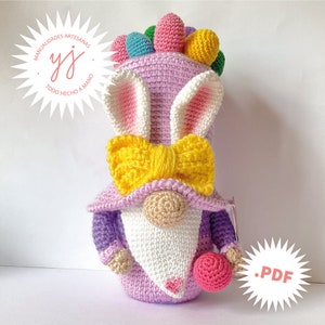 May include: A crocheted Easter gnome wearing a purple hat, white beard, and yellow bow. The gnome is holding a pink ball and has a pink and white bunny ear hat with a rainbow of crocheted balls on top.