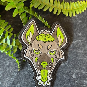 以下が含まれることがあります： 脳のデザインが施された、様式化されたハイエナの頭部が特徴の刺繍パッチ。パッチは黒い縁取りで、タン、ブラウン、ライムグリーンのアクセントが付いています。ハイエナの舌が出ています。パッチは濃い灰色の表面にあります。