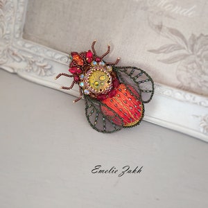 May include: A handcrafted, beaded insect brooch in shades of red, orange, and gold. The detailed design features a central yellow and iridescent section, surrounded by red gemstones and intricate beadwork. The wings are outlined with green beads. The artist's name, Emelie Zakh, is at the bottom.