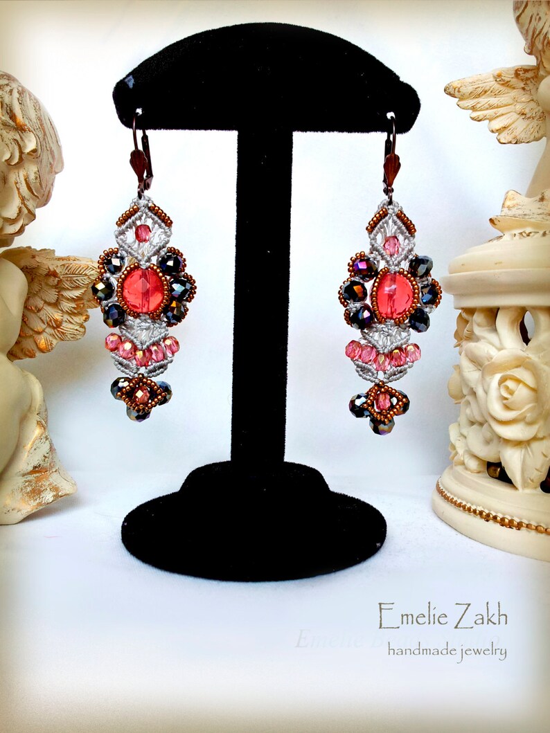May include: A pair of silver and pink beaded earrings with a pink crystal in the center of each earring. The earrings are hanging from a black stand. The text "Emelie Zakh handmade jewelry" is visible at the bottom of the image.
