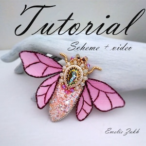 Könnte beinhalten: Eine rosa und goldene Perlen-Insektenbrosche mit einem großen, schillernden grünen Edelstein in der Mitte. Die Brosche befindet sich auf einer weißen Oberfläche mit dem Text "Tutorial Schema + Video" darüber und "Emelie Zakk" darunter.