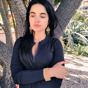 May include: A woman wearing a black long-sleeved shirt with a zipper. She is wearing gold earrings and a gold bracelet. The woman is standing in front of a tree.