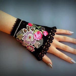 Puede incluir: Puño de encaje negro para la muñeca con detalles florales bordados en rosa, morado y verde. El puño está adornado con pequeñas cuentas brillantes.