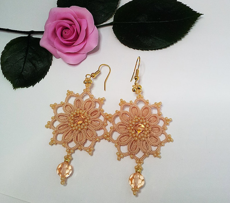 Pode incluir: Um par de brincos de croch&ecirc; cor p&ecirc;ssego com ganchos dourados e um pingente de cristal transparente. Os brincos t&ecirc;m a forma de uma flor com um centro de contas.