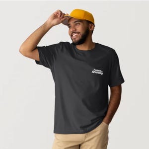 Może przedstawiać: Czarny t-shirt z białym napisem "Tuned Jeremy" na lewej piersi. Osoba na zdjęciu ma na sobie żółtą czapkę z daszkiem i spodnie khaki.