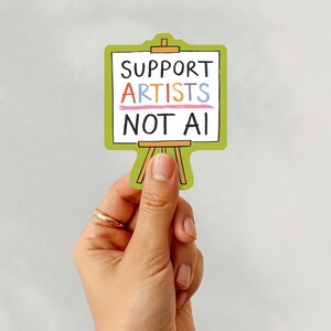 A white woman's hand holding a paper sticker illustrated by Hayley Wells. The design features an artist's canvas painted with the words "support artists, not AI". The sticker has a green background.