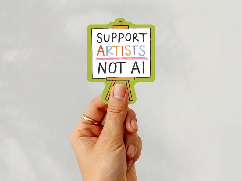 A white woman's hand holding a paper sticker illustrated by Hayley Wells. The design features an artist's canvas painted with the words "support artists, not AI". The sticker has a green background.