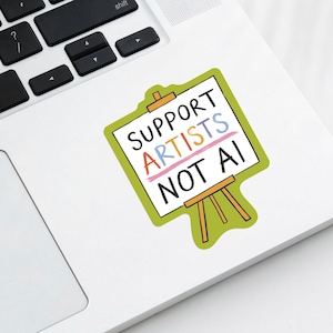 The corner of a modern laptop with a paper sticker illustrated by Hayley Wells. The design features an artist's canvas painted with the words "support artists, not AI". The sticker has a green background.