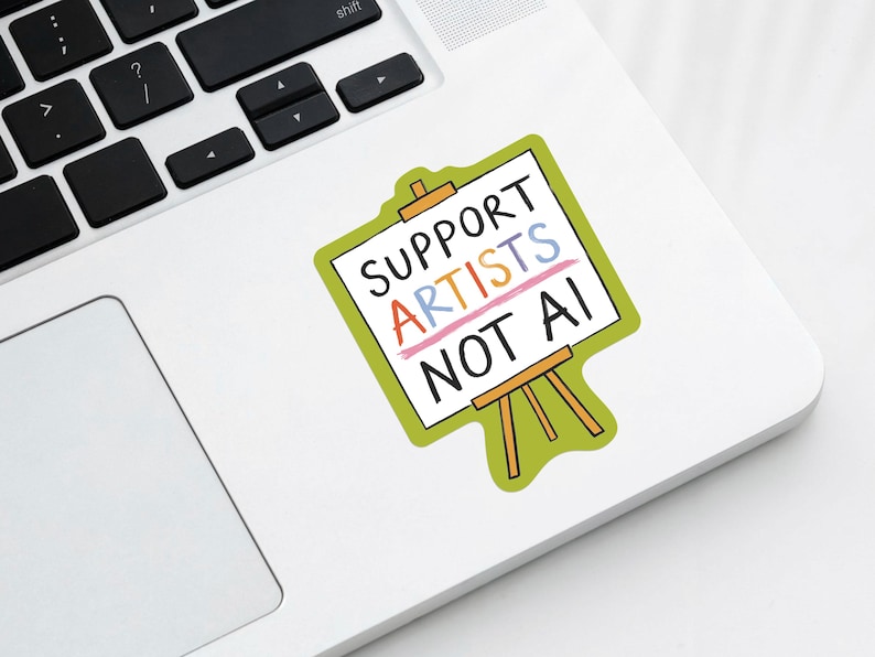 The corner of a modern laptop with a paper sticker illustrated by Hayley Wells. The design features an artist's canvas painted with the words "support artists, not AI". The sticker has a green background.