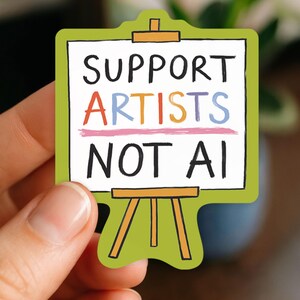 A white woman's hand holding a paper sticker illustrated by Hayley Wells. The design features an artist's canvas painted with the words "support artists, not AI". The sticker has a green background.