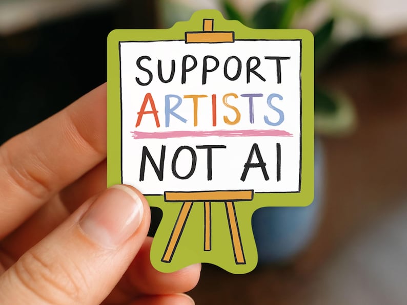 A white woman's hand holding a paper sticker illustrated by Hayley Wells. The design features an artist's canvas painted with the words "support artists, not AI". The sticker has a green background.