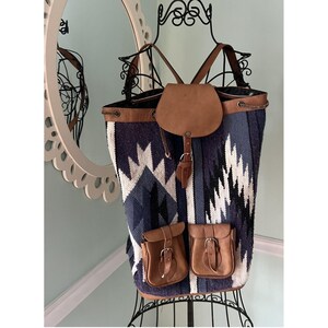 May include: A large backpack with a southwestern pattern in blue, black, and white. The bag has brown leather accents, including a flap closure and two front pockets. The straps are also brown leather.